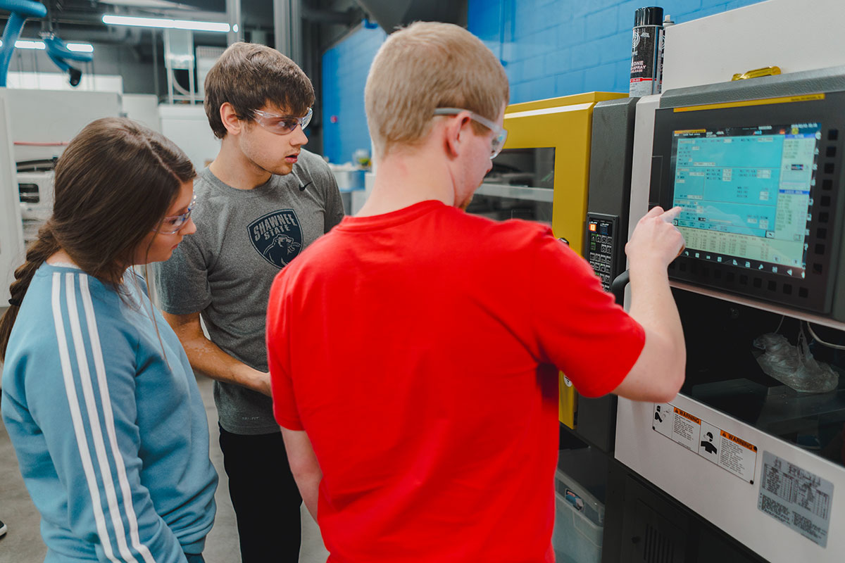 Three Shawnee State University Plastics Engineering Technology students wearing safety glasses work together at an injection molding machine in the Advanced Manufacturing Center.