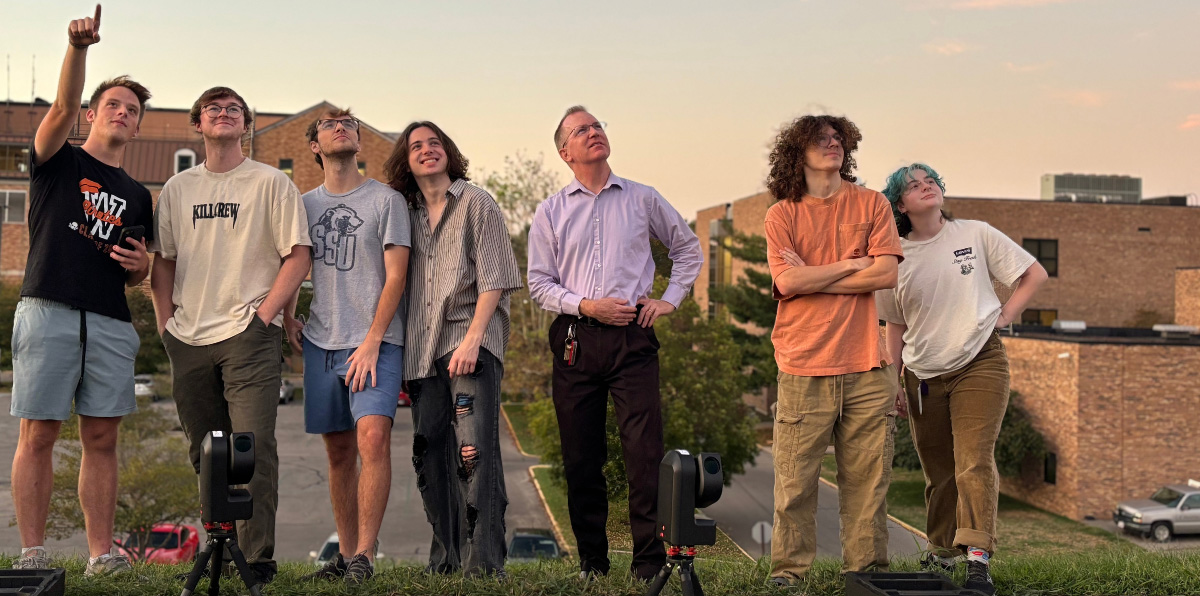 Group of seven people standing outdoors at sunset, looking upward together in front of SSU campus buildings