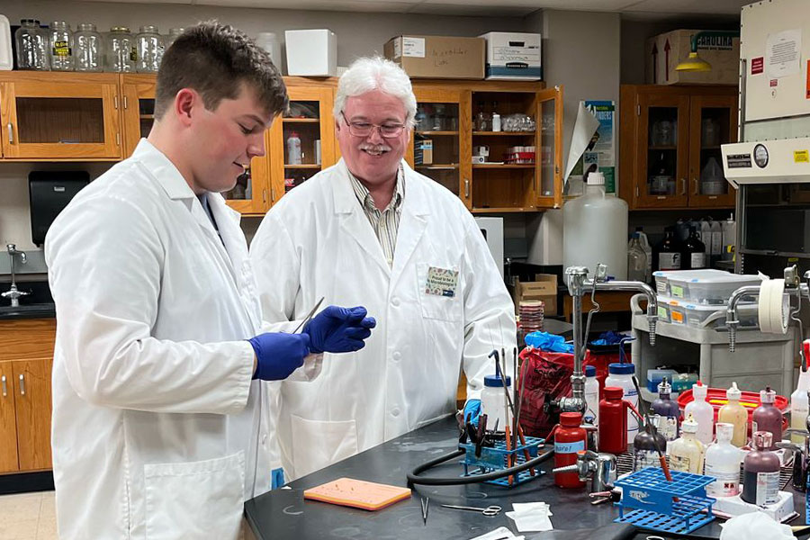 Two people in lab coats working together at a laboratory bench with various bottles and equipment.