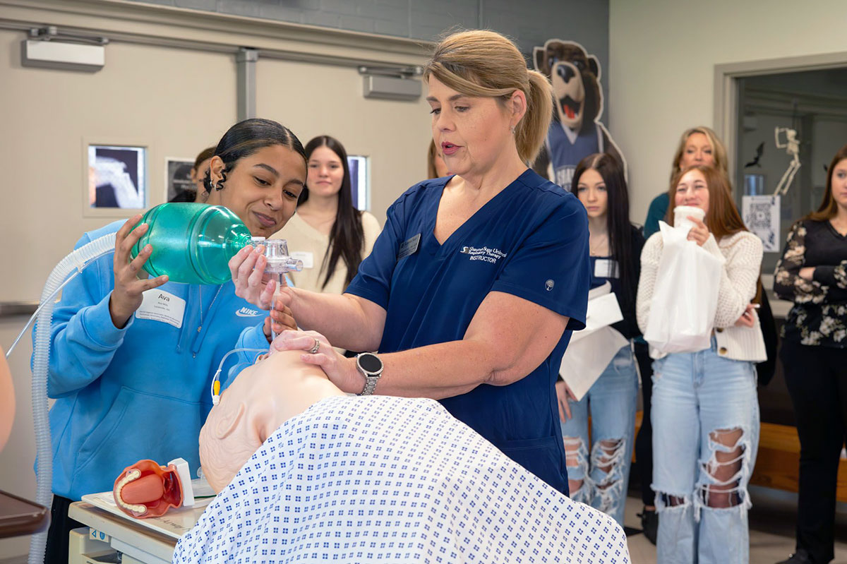 instructor demonstrates bag-valve-mask ventilation on a medical mannequin while students observe.