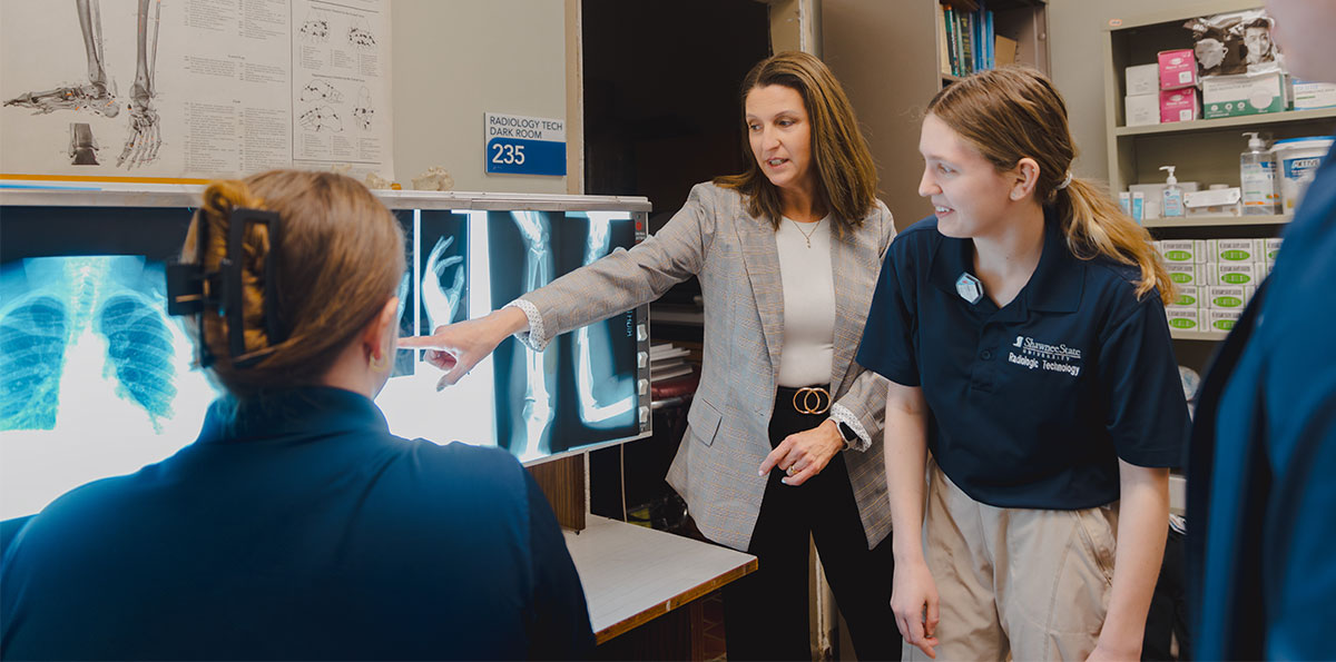 Instructor points to X-ray images while radiologic technology students review scans in a lab.