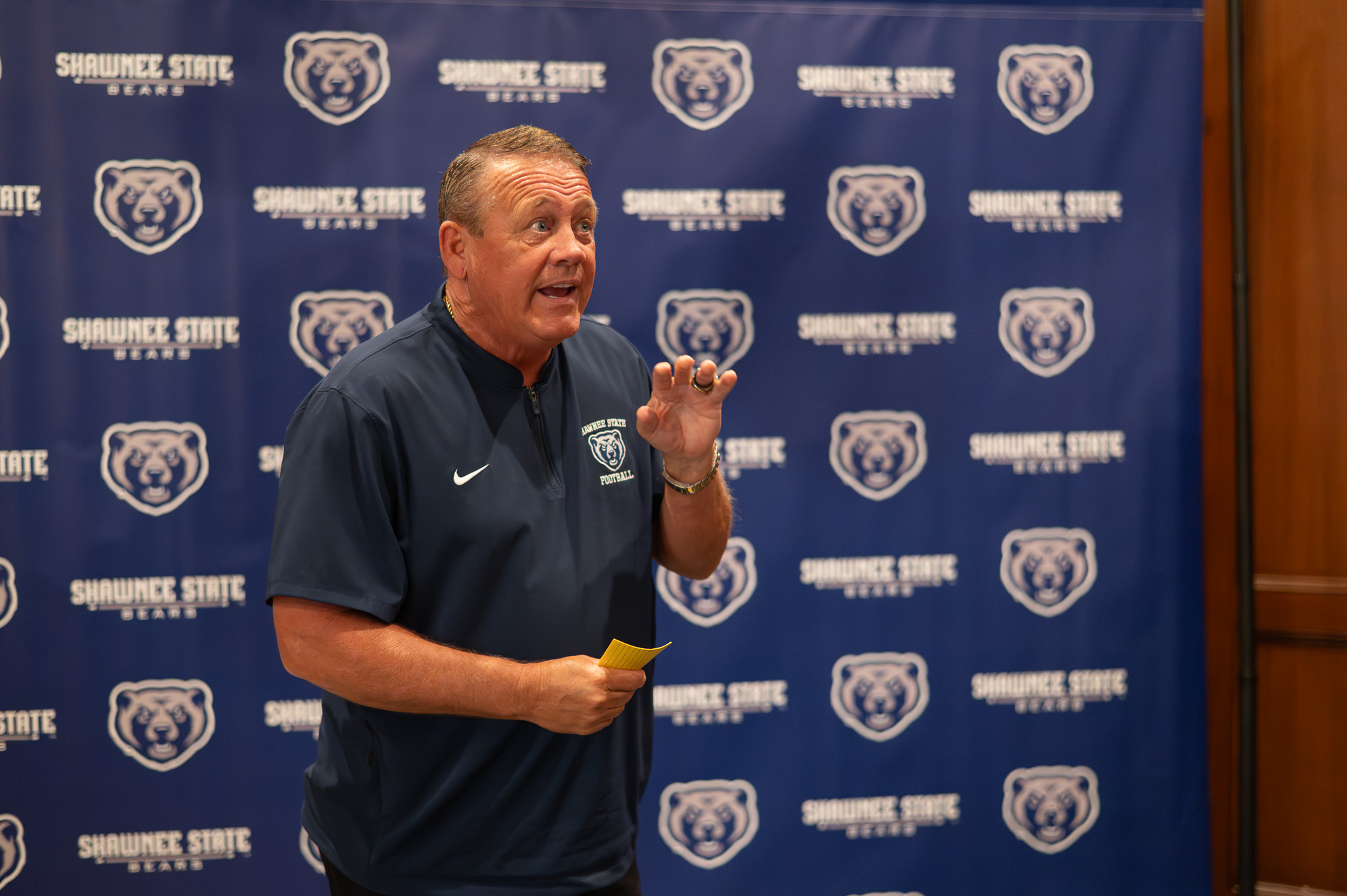 A man wearing a navy Shawnee State Bears football pullover speaks in front of a blue backdrop covered with Shawnee State Bears logos, gesturing with one hand while holding a small yellow note card.