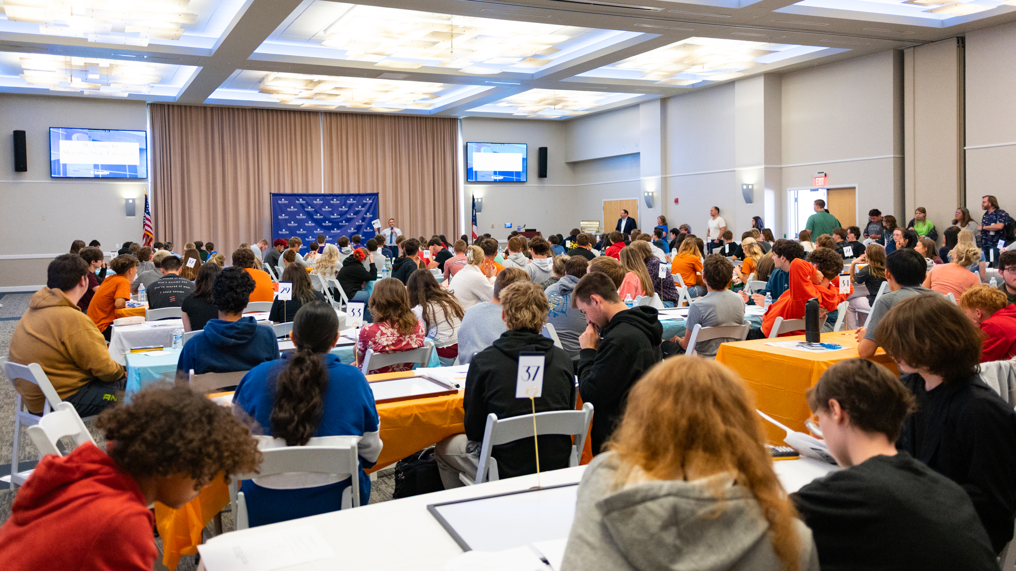 Students seated at numbered tables in a large banquet hall compete in the 2026 SUMS Math Competition at Shawnee State University, with a presenter at the front of the room near a blue branded backdrop.