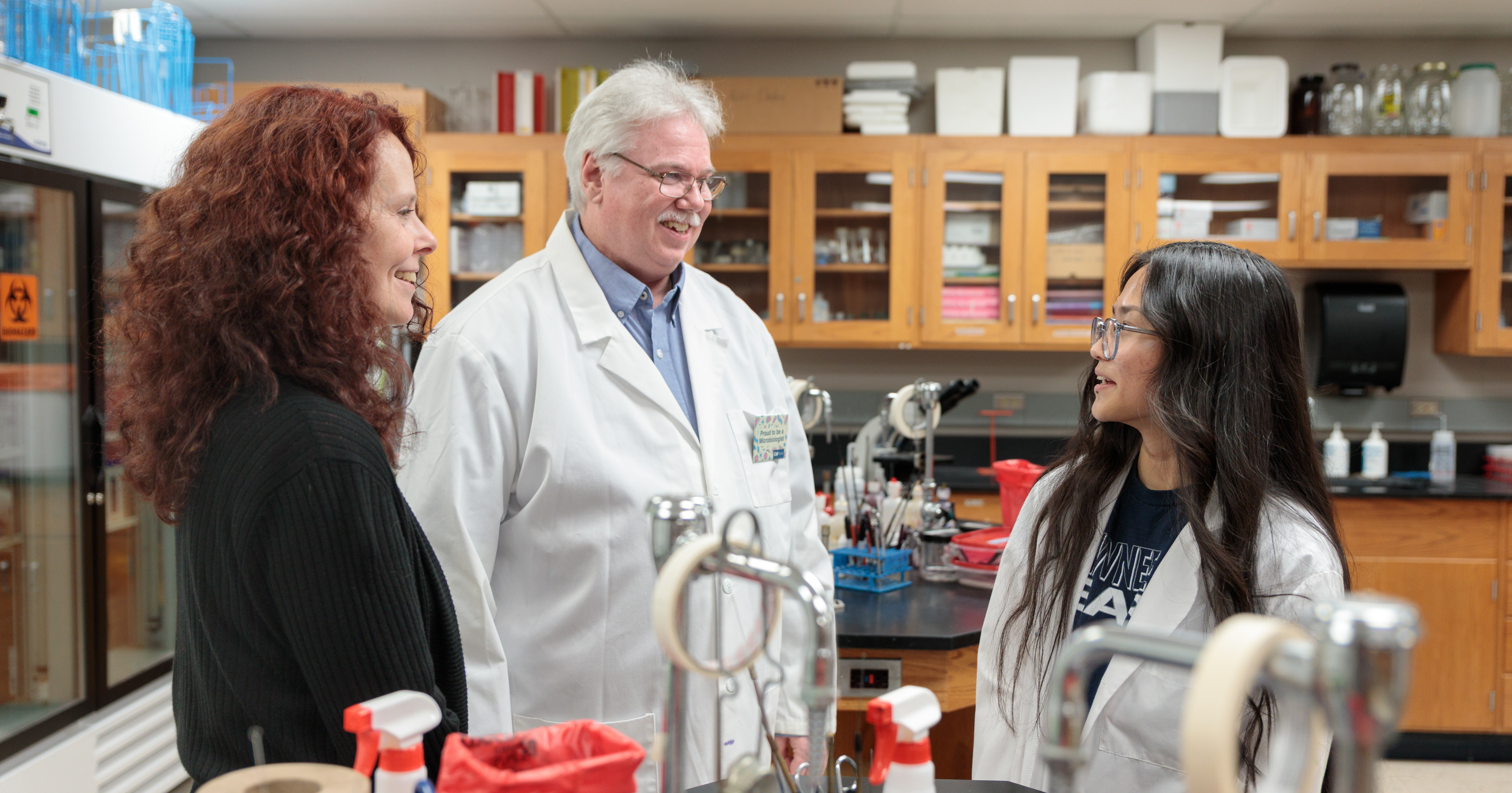 Three people chatting in a laboratory, with two wearing lab coats and scientific equipment visible in the background.