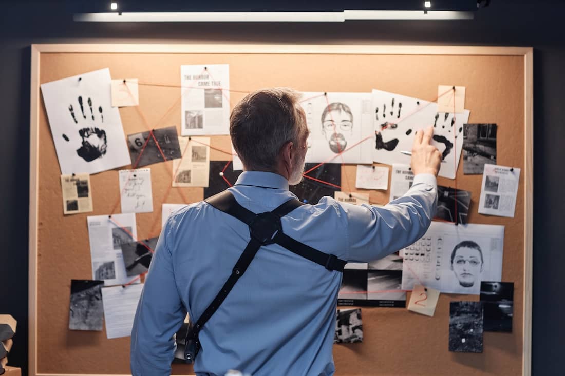 Back view of senior detective standing by evidence board and studying leads in investigation.