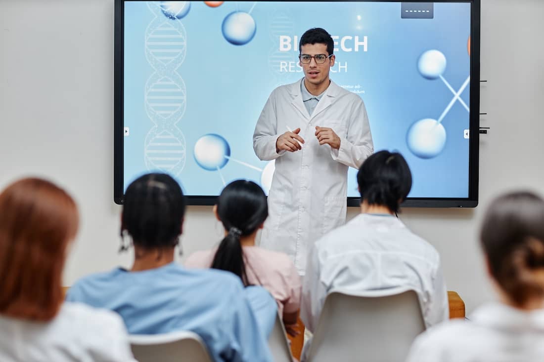 Portrait of young medical professional giving lecture or presentation standing by screen at seminar and speaking to audience.