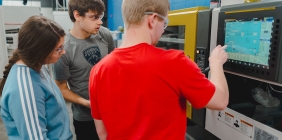 Three Shawnee State University Plastics Engineering Technology students wearing safety glasses work together at an injection molding machine in the Advanced Manufacturing Center.