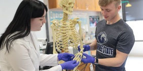 Two students examine a human skeleton model in a lab while wearing gloves.