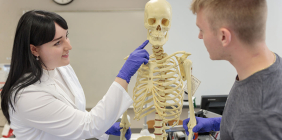 Two students examine a human skeleton model in a lab while wearing gloves.
