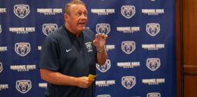 A man wearing a navy Shawnee State Bears football pullover speaks in front of a blue backdrop covered with Shawnee State Bears logos, gesturing with one hand while holding a small yellow note card.