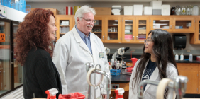 Three people chatting in a laboratory, with two wearing lab coats and scientific equipment visible in the background.