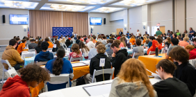 Students seated at numbered tables in a large banquet hall compete in the 2026 SUMS Math Competition at Shawnee State University, with a presenter at the front of the room near a blue branded backdrop.