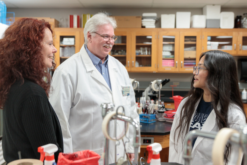 Three people chatting in a laboratory, with two wearing lab coats and scientific equipment visible in the background.
